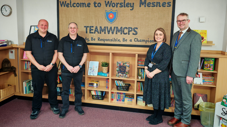 A group of four people stand smiling in front of a bookshelf. A banner above reads "Welcome to Worsley Mesnes." The setting is warm and inviting.